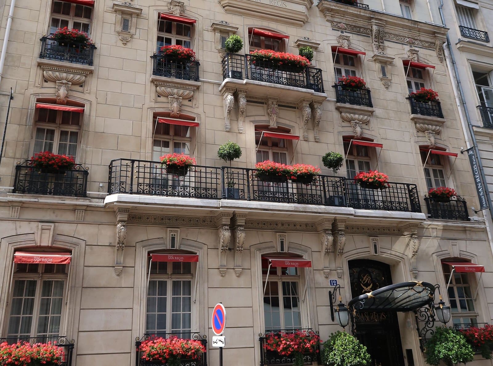 Photograph of the stone front of the Hôtel San Régis on rue Jean-Goujon, with red awnings lettered San Régis over the windows, window boxes of red geraniums, a long iron balcony, and a glass-and-iron canopy over the entrance at number 12.