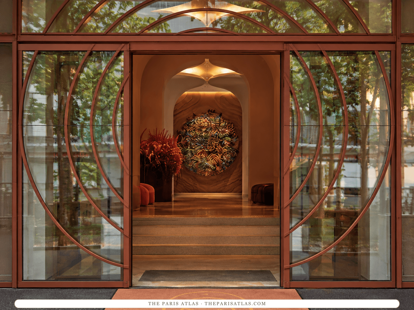 AI-generated illustration of the entrance to SO/ Paris: tall glass doors in curved copper-colored frames opening onto a warm-lit hall with an arched ceiling and a round woven artwork on the far wall.