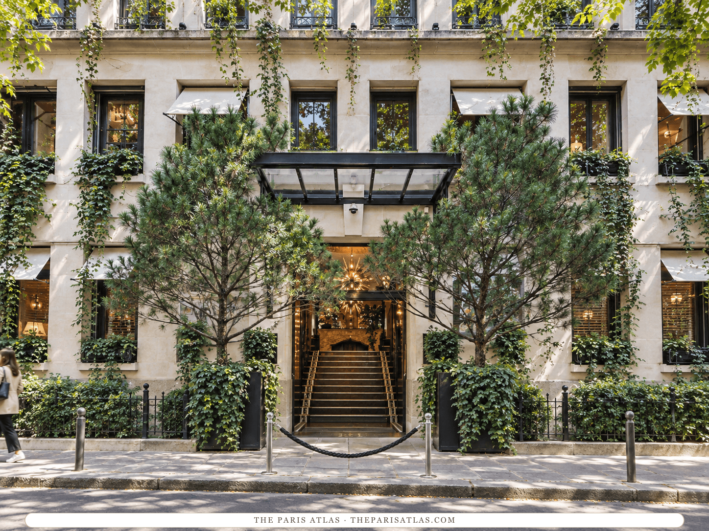 Reference-based illustration of the pale stone front of The One Alma Paris: a flat black-and-glass canopy over a staircase leading up into a lit lobby, two pine trees in planters, ivy on the walls and a rope barrier on the pavement.