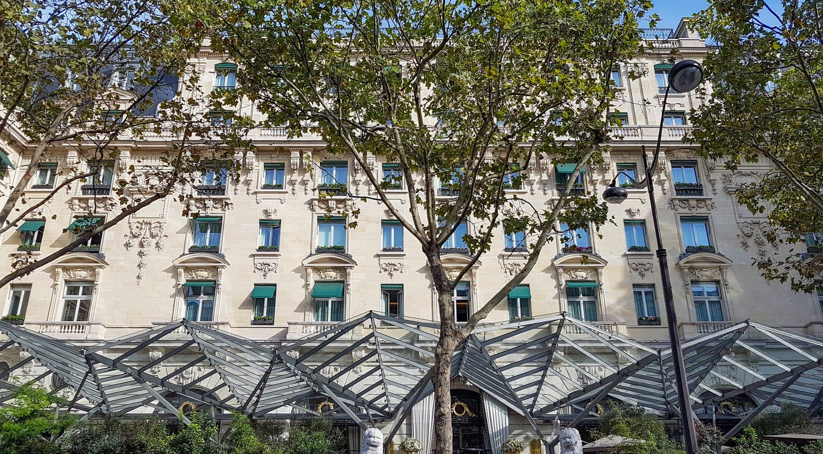 Photograph of the pale carved stone front of The Peninsula Paris on avenue Kléber in sunshine, five storeys with green awnings, behind plane trees and above the angular glass-and-steel canopy of the hotel's terrace.