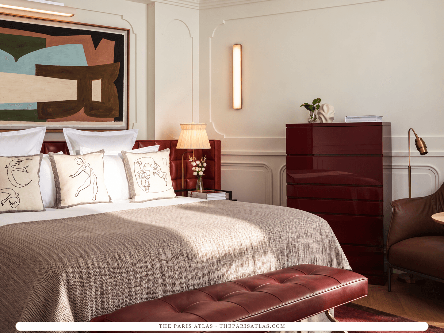 AI-generated illustration of a bedroom at Villa-des-Prés: a bed with cushions printed with line drawings, a burgundy headboard and bench, a lacquered red chest of drawers, a brass wall light and a large abstract painting.
