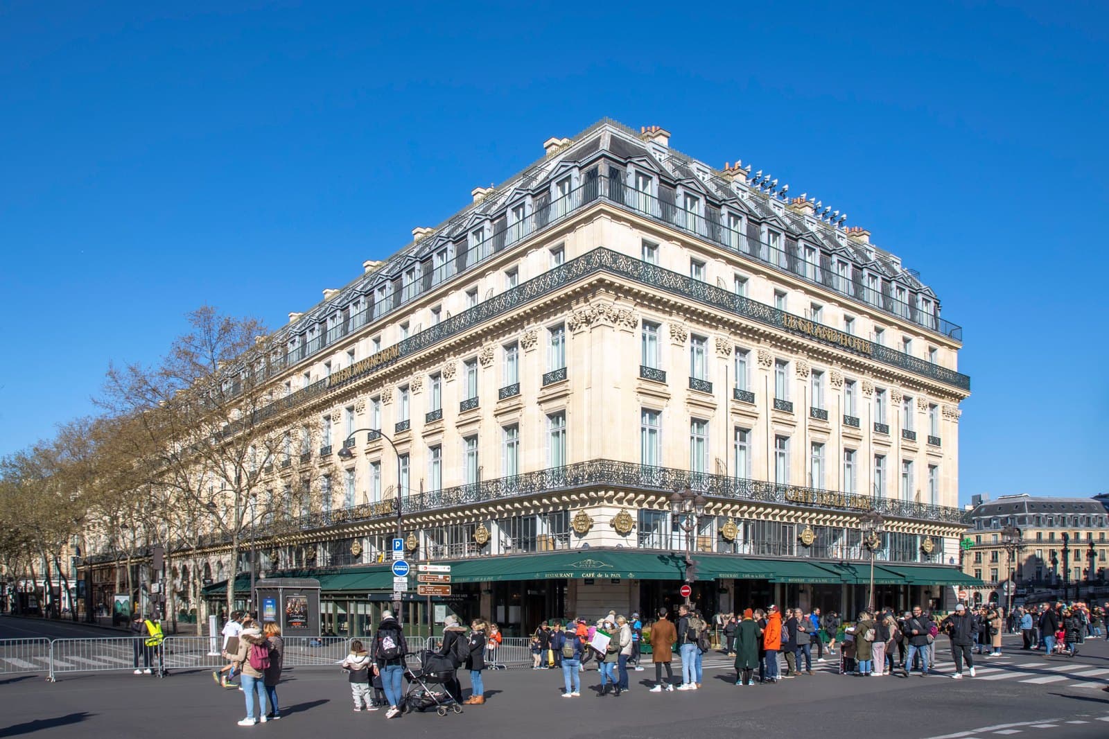 Photograph of the long stone façade of the Grand Hôtel on place de l'Opéra under a blue sky, with the green awnings of the Café de la Paix along the ground floor and people crossing the square.