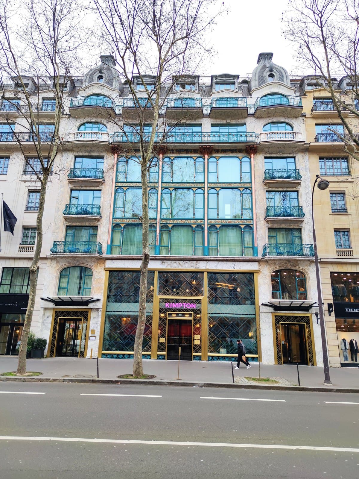 Photograph of the Art Nouveau façade of Kimpton St Honoré on boulevard des Capucines, with turquoise iron-and-glass bays over three storeys, a pink Kimpton sign above the central entrance and bare trees in front.