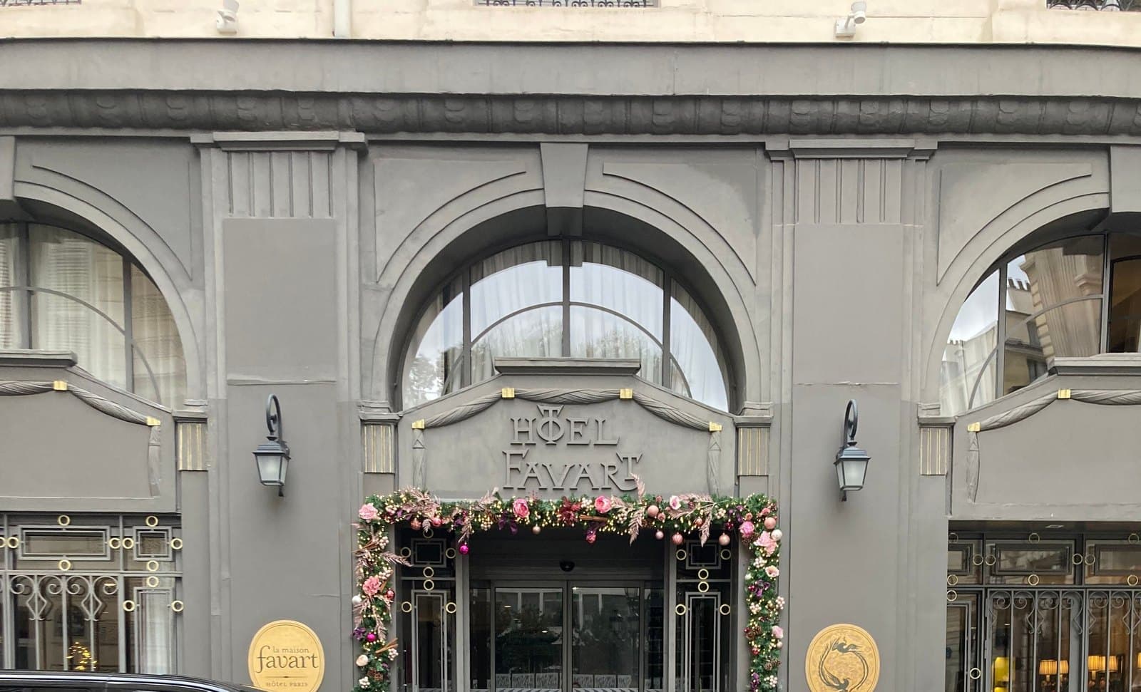 Photograph of the grey-painted ground floor of La Maison Favart on rue de Marivaux, with arched windows, Hôtel Favart lettered above the glass entrance, a garland of pink and white flowers around the door, two lanterns and two round gold plaques.