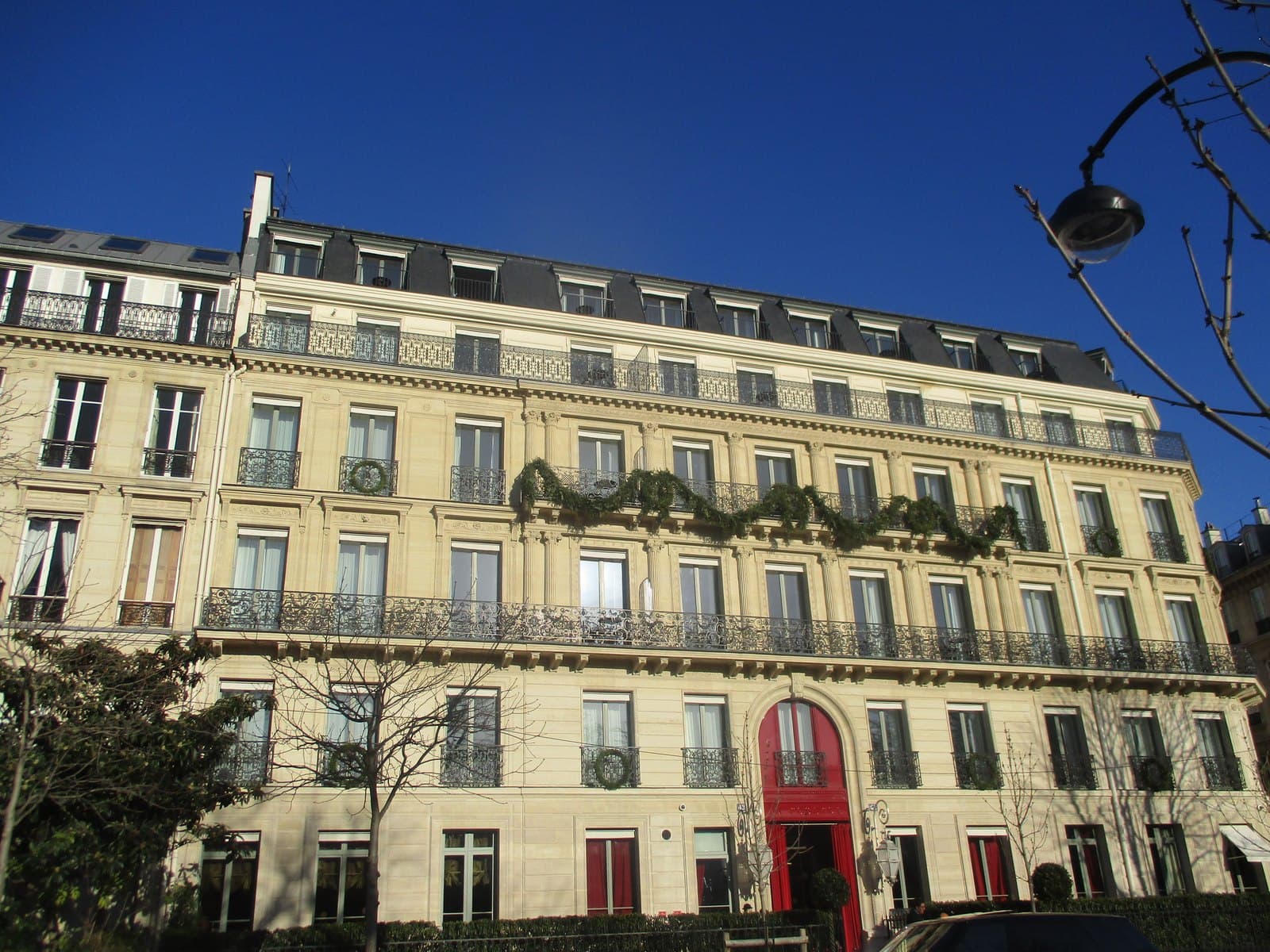 Photograph of the cream stone front of La Réserve on avenue Gabriel in winter sun, with three floors of wrought-iron balconies hung with Christmas garlands, a set-back top floor with slate dormers and an arched red lacquered entrance.