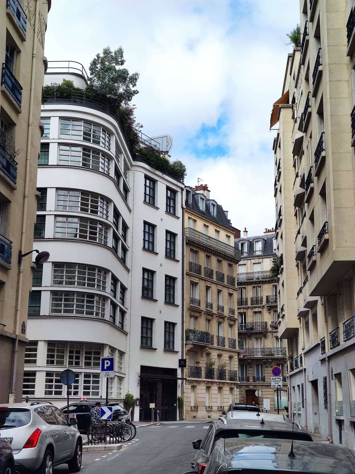 Photograph of Le Cinq Codet, a white building with a rounded corner, wide bands of small-paned windows and trees on its roof terrace, with a dark canopy lettered Le Cinq Codet over the entrance, between stone apartment buildings on a narrow street with parked cars.