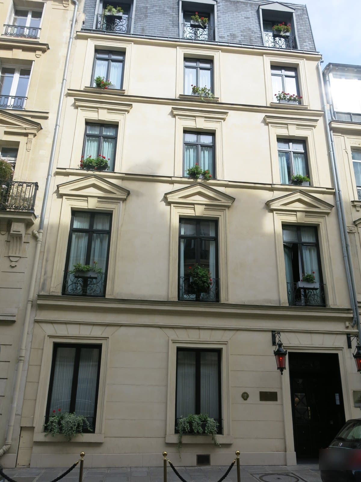 Photograph of the cream-rendered five-storey front of Maison Souquet, three windows wide, with pedimented first-floor windows, iron window guards, red geraniums in window boxes, a mansard roof, and two red lanterns on iron brackets beside a dark glazed entrance door, with gold posts and a rope on the pavement.