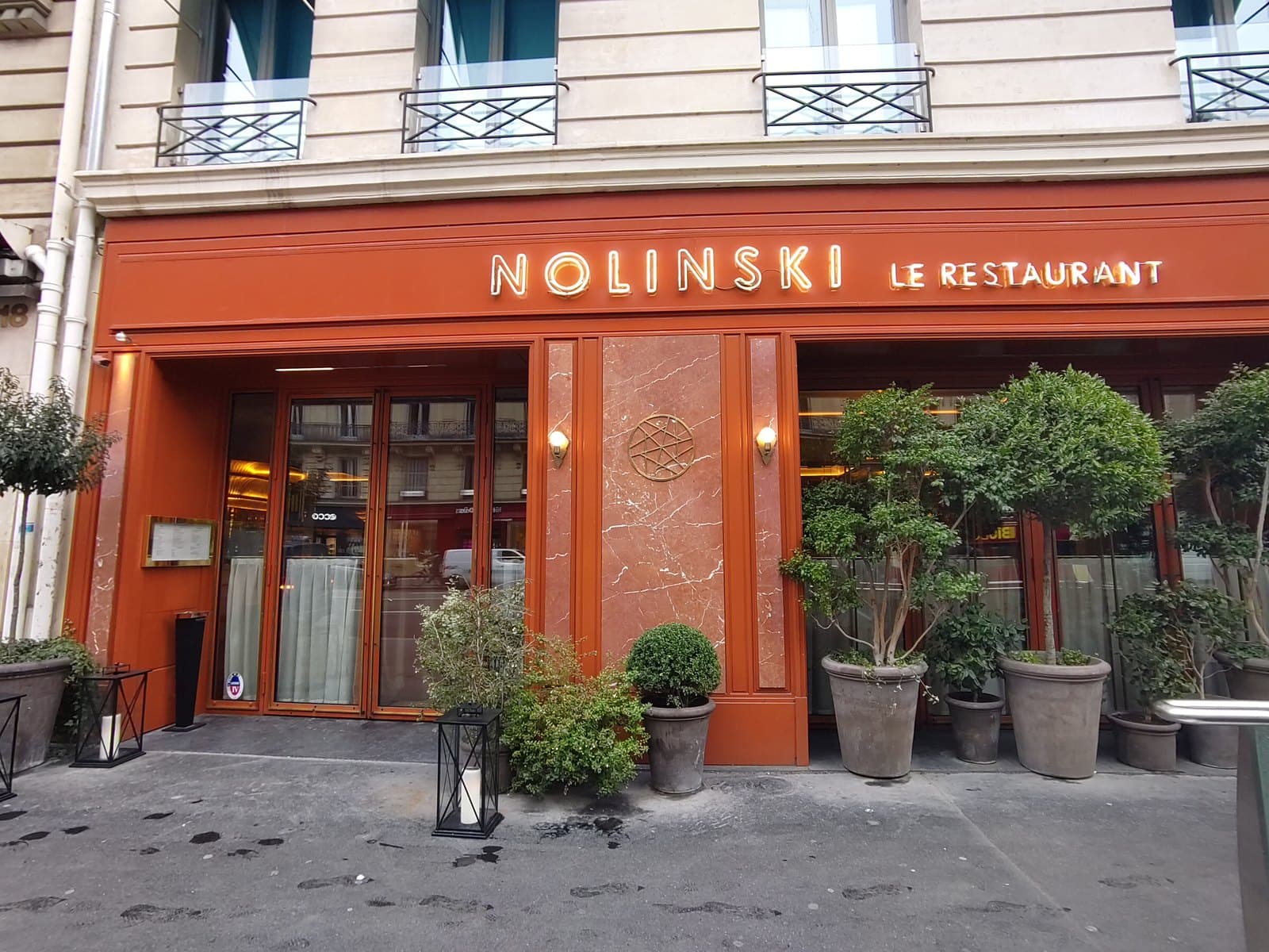 Photograph of the Nolinski ground-floor frontage on avenue de l'Opéra: red marble panels, gold letters reading Nolinski Le Restaurant, and potted trees on the pavement.