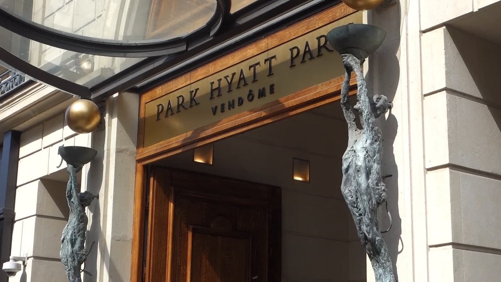 Photograph looking up at the hotel entrance on rue de la Paix: a gilded sign reading PARK HYATT PARIS VENDÔME above wooden doors, between two tall bronze figures holding up bowl-shaped lamps, under a glass canopy.