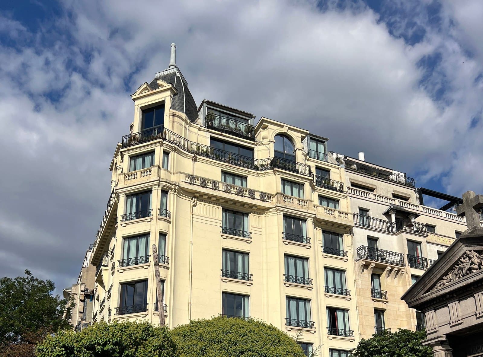 Photograph of the cream stone front of Terrass" Hôtel in sunshine under a cloudy sky, with its slate-domed corner pavilion, rows of iron balconies and a glazed roof terrace, above a line of trees.