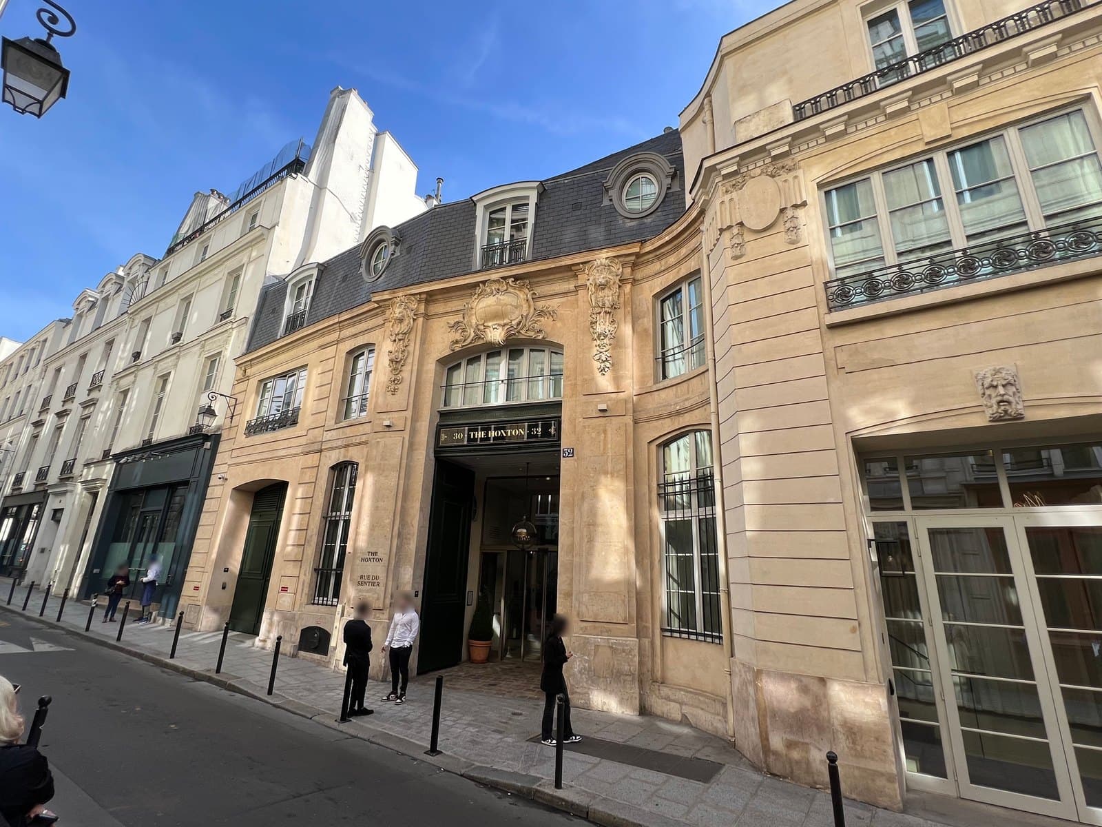 Photograph of The Hoxton's carved stone front on rue du Sentier in sunshine, with a slate mansard roof, a central carriage arch under a sign reading 30 The Hoxton 32, and a stone plaque reading The Hoxton, rue du Sentier.
