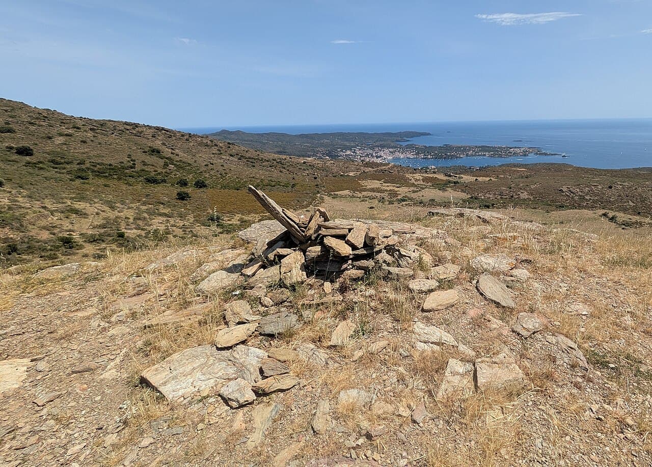 Wide view from Puig de sa Cruïlla across dry rocky ground to the small white town and blue bay of Cadaqués