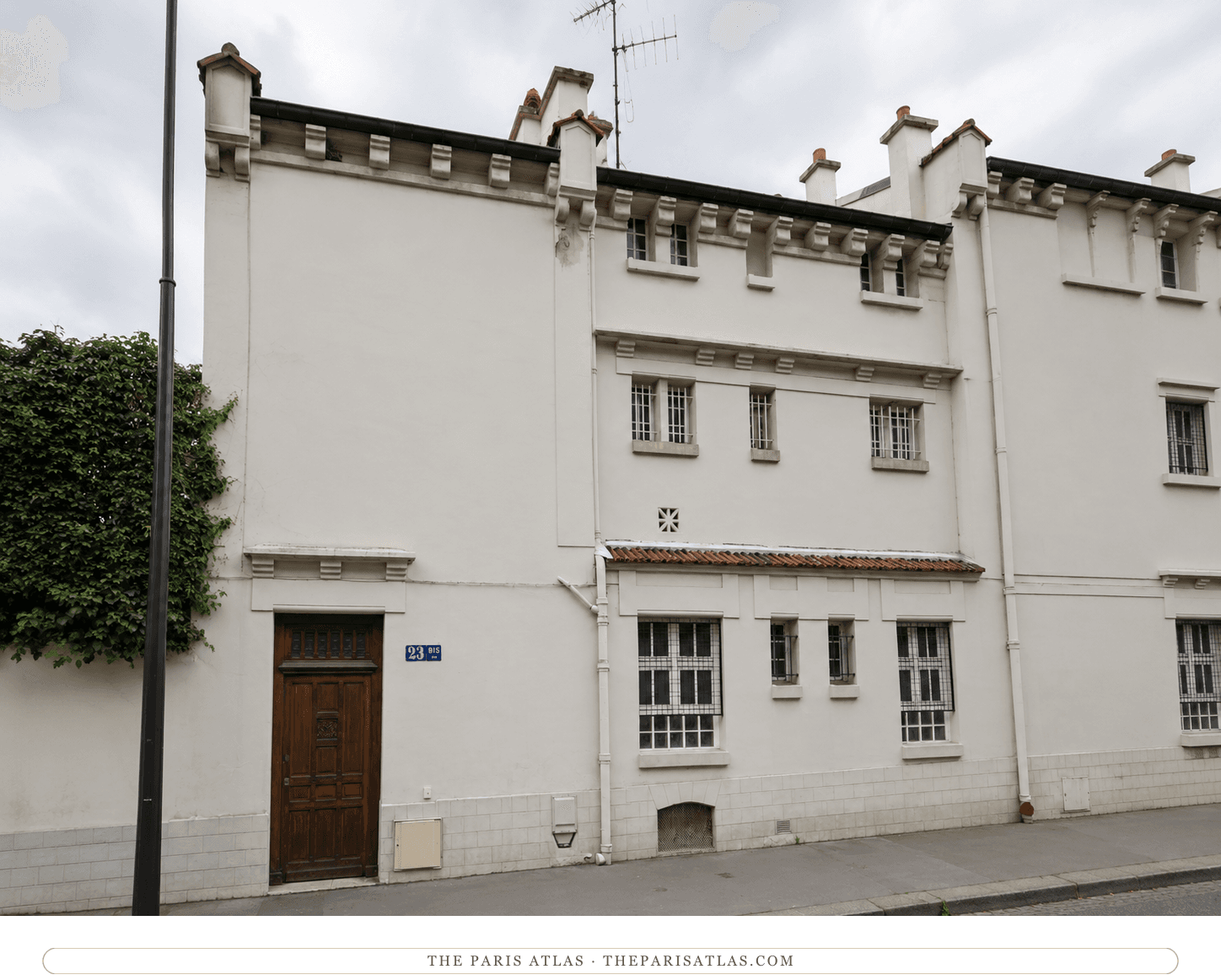 The same rue Jean-Dolent house front in color, cream walls under a grey sky, red-tiled ledges and ivy on the wall beside the lamp post