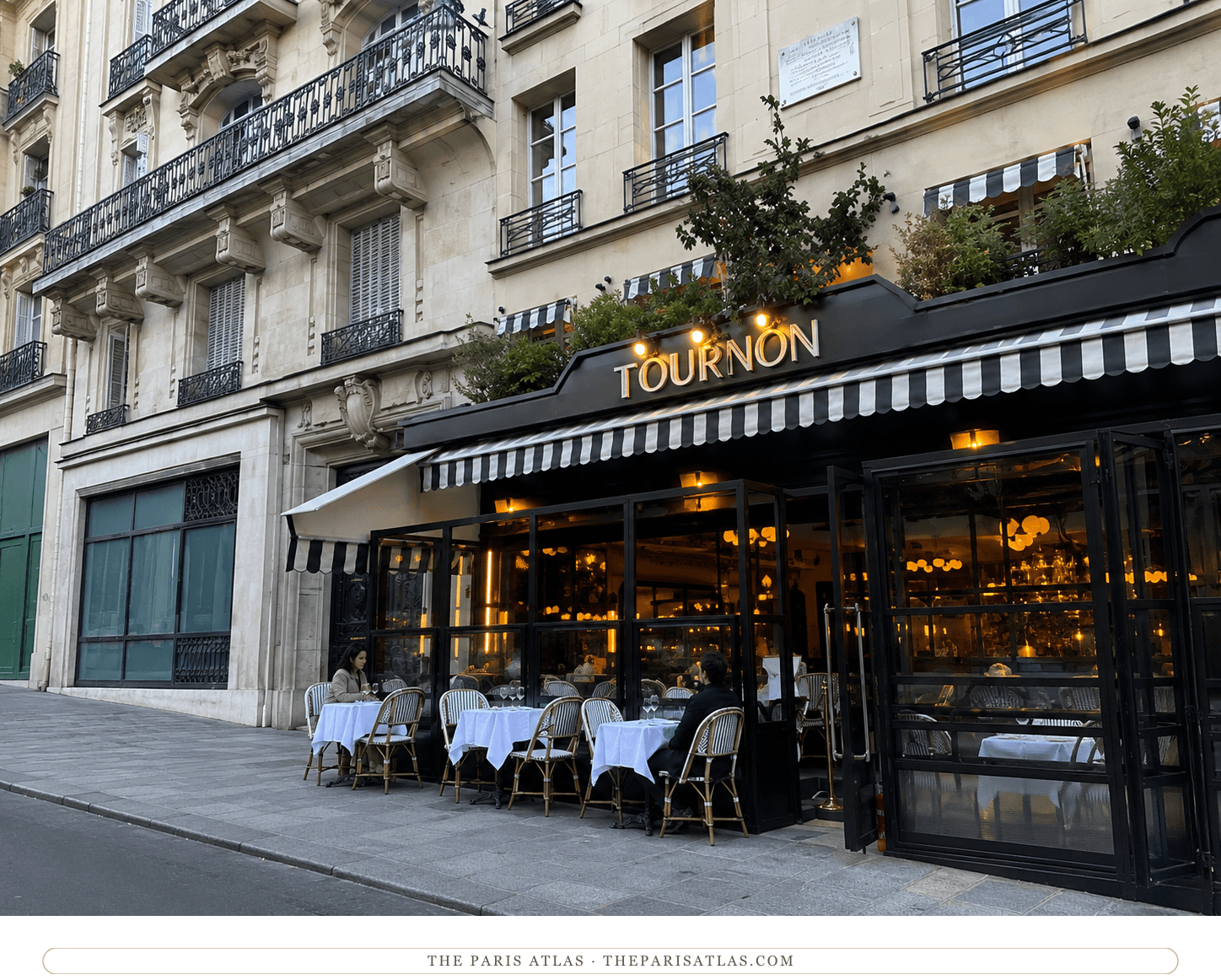 Le Tournon today, black and white striped awning over terrace tables, warmly lit dining room behind the glass