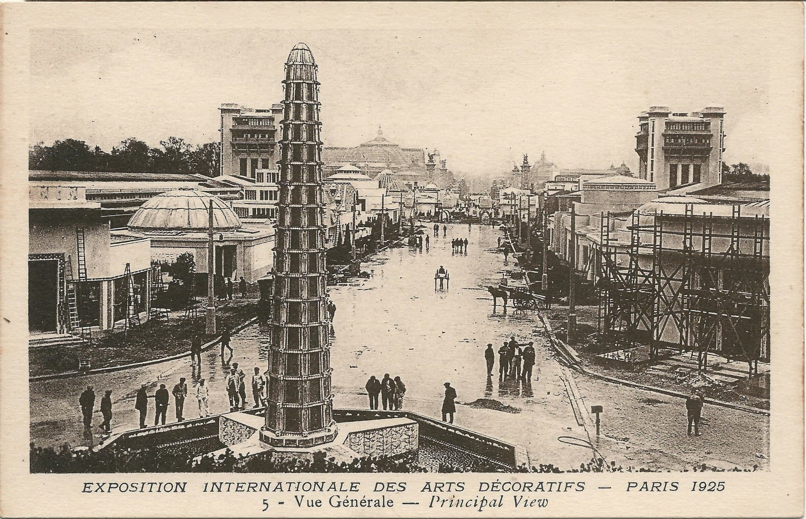General view along the main avenue of the 1925 Paris exhibition, a tall tiered tower in the foreground, domed and stepped pavilions on either side, workmen and scaffolding still in place along the wet path