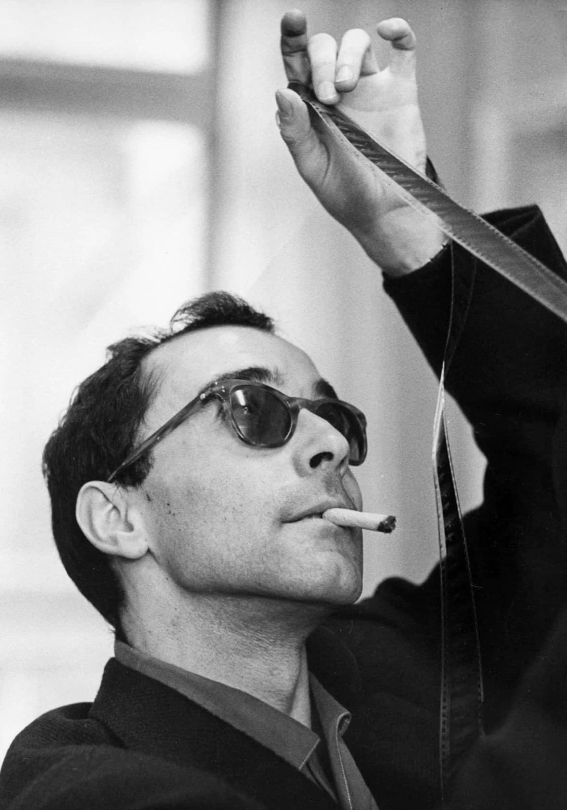 Young Jean-Luc Godard in dark glasses, cigarette in his mouth, holding a strip of film up to the light with both hands