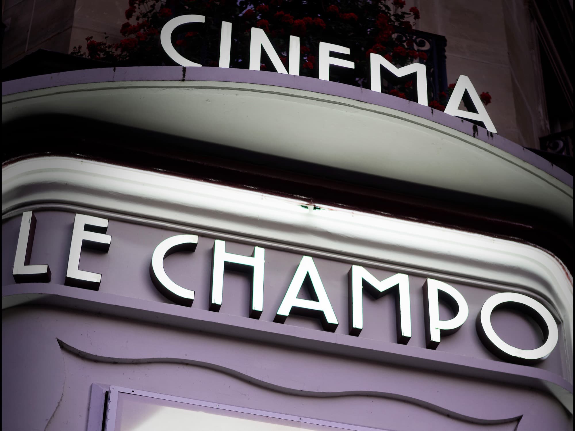 The lit night marquee of Le Champo cinema in the Latin Quarter: CINEMA above, LE CHAMPO in glowing deco letters, red geraniums on the balcony above