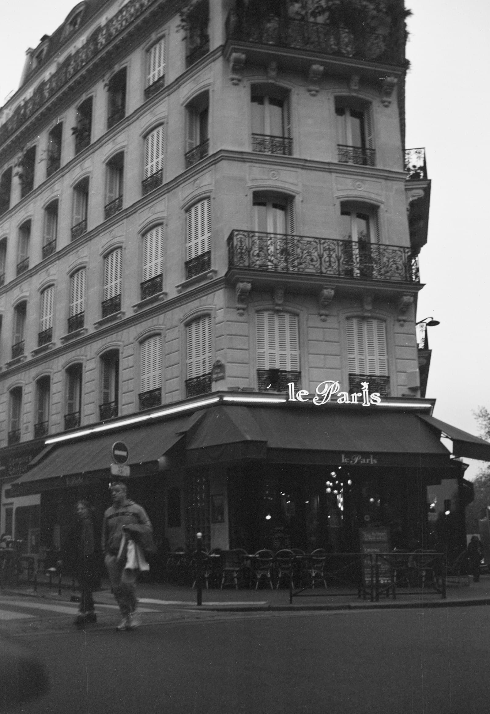 Black-and-white film photograph of a Paris corner café, its awning glowing over empty terrace chairs