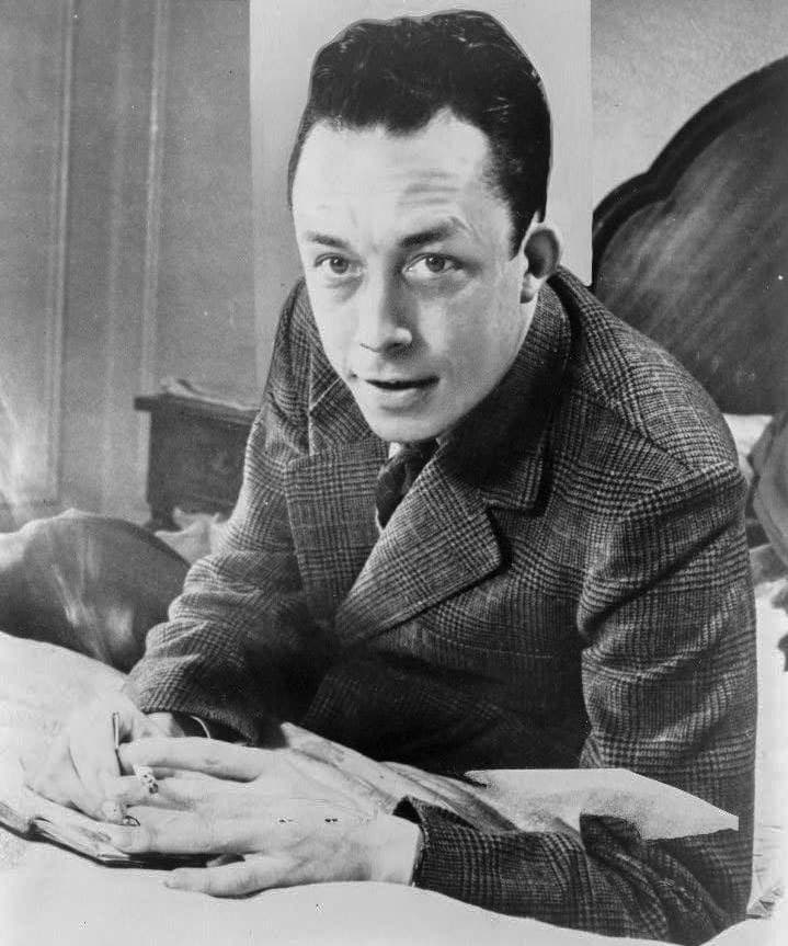Albert Camus seated at a desk in 1957, turned in profile with a cigarette in his hand.