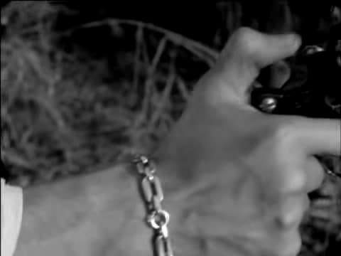 A hand wearing a chain bracelet aims a revolver out of a moving car at the roadside, in Breathless