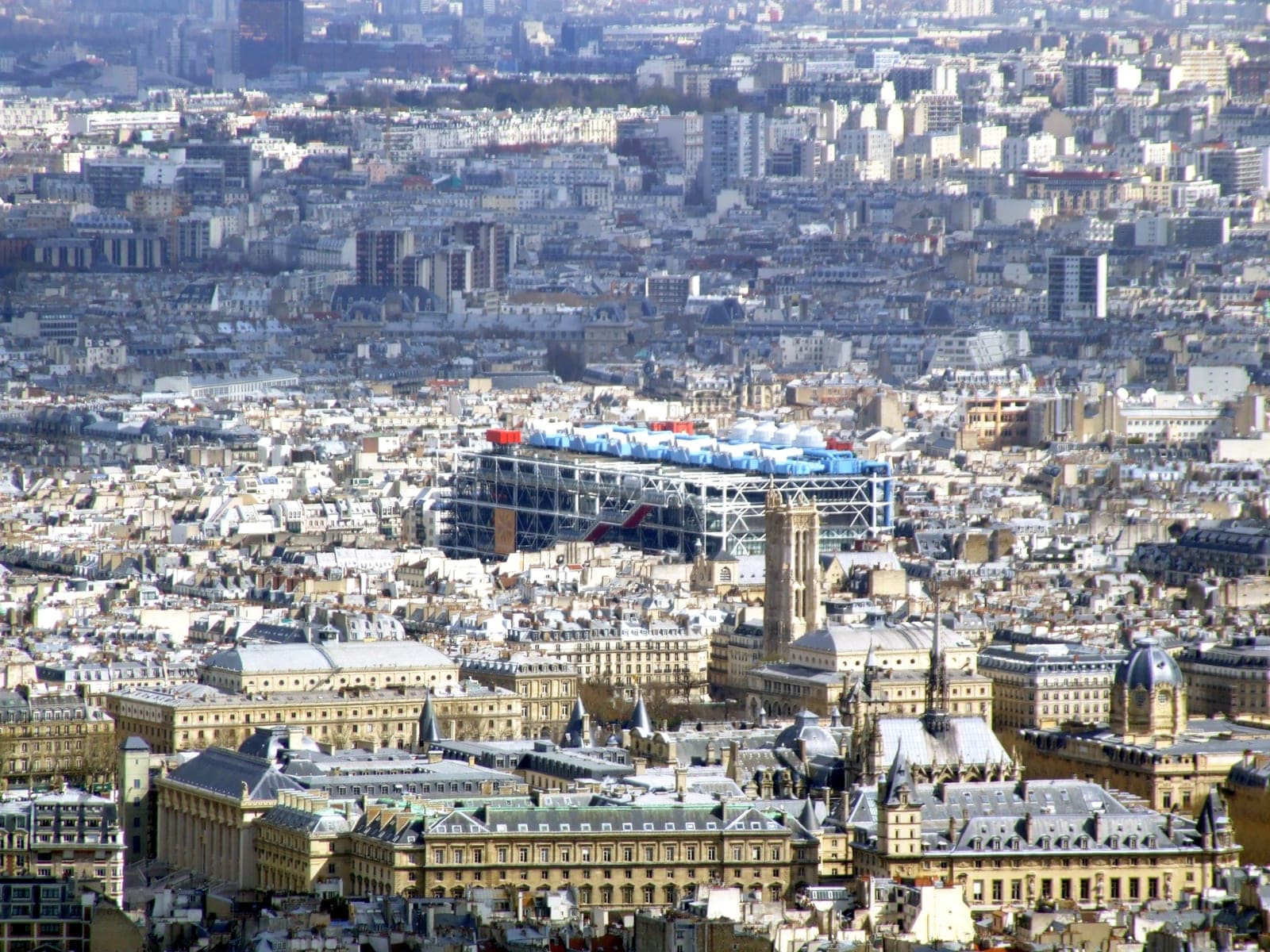 The Centre Pompidou's blue-and-white shell rising above the Paris roofscape, seen from the Tour Montparnasse, with the Tour Saint-Jacques to its right