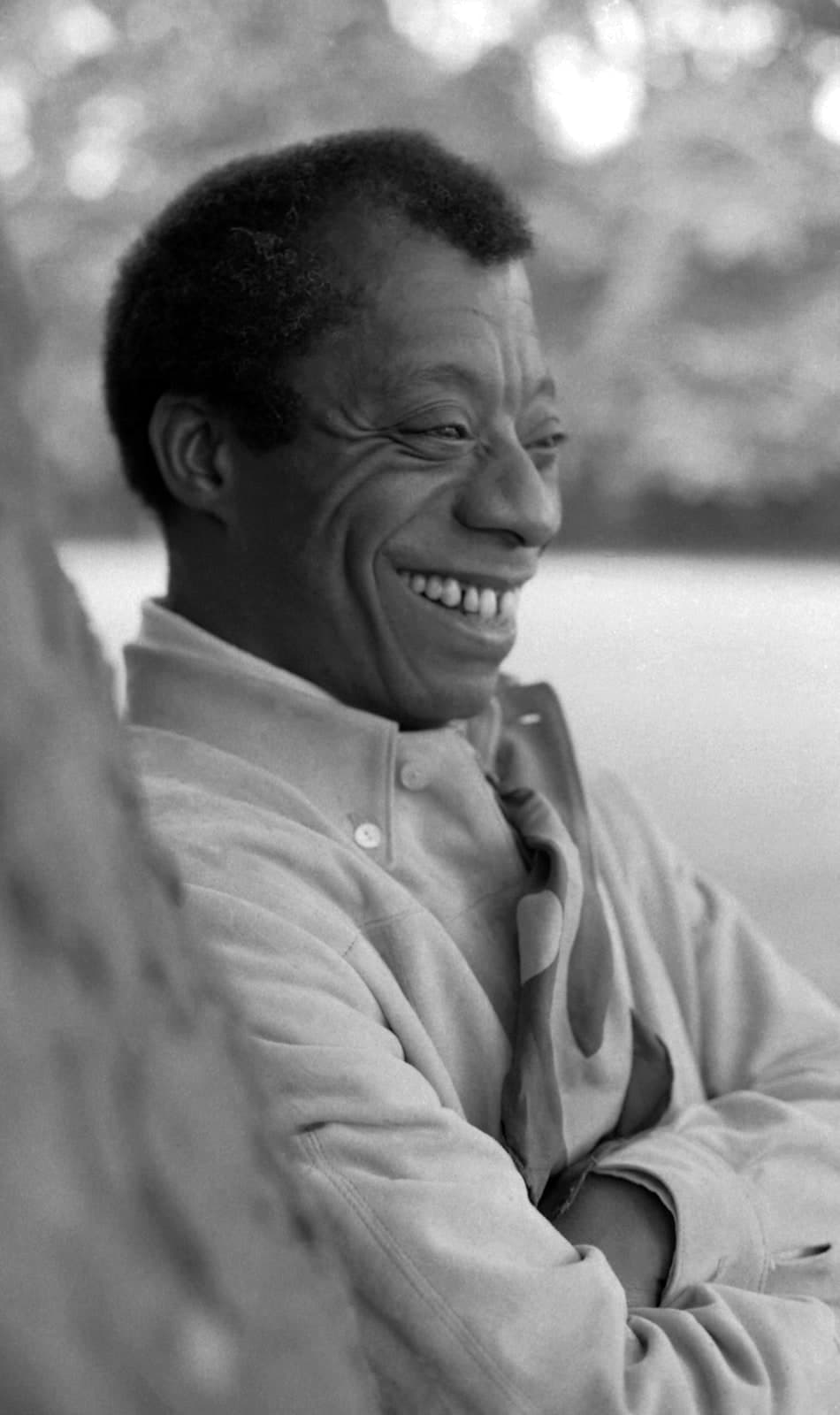 James Baldwin laughing in profile, wearing a pale jacket and scarf, photographed outdoors in Hyde Park, London, in 1969