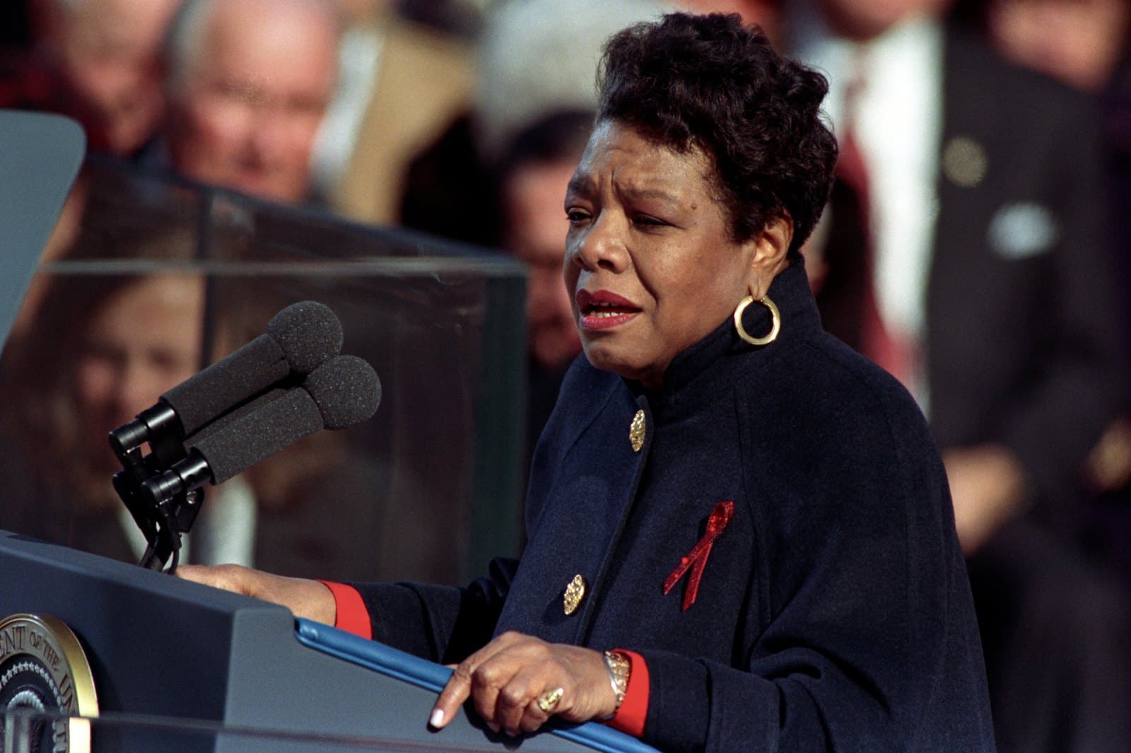 Maya Angelou reciting from a lectern bearing the presidential seal at an outdoor ceremony, wearing a dark coat and a red ribbon, microphones before her and dignitaries behind