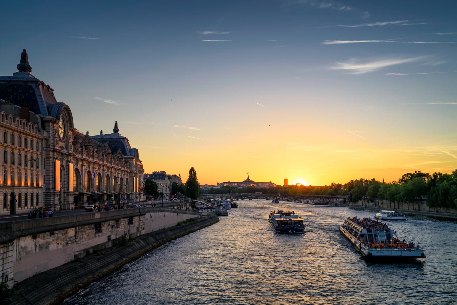The Musée d'Orsay at sunset beside the Seine, its former station clock and stone river façade glowing above passing boats